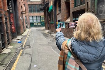 A lady in grey coat, taking a picture with a phone of a photograph in an alley between two red brick buildings