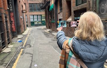 A lady in grey coat, taking a picture with a phone of a photograph in an alley between two red brick buildings