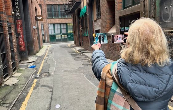 A lady in grey coat, taking a picture with a phone of a photograph in an alley between two red brick buildings