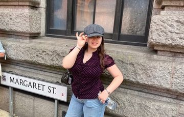 A lady in dark red top wearing a cloth cap next to to a road sign saying Margaret St