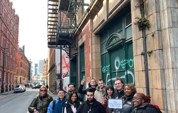 A large group of people standing on a pavement holding a film clapper and a sign The Locationist Film Tour