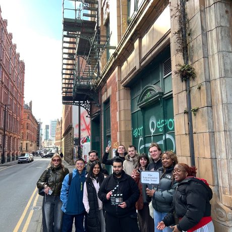 A large group of people standing on a pavement holding a film clapper and a sign The Locationist Film Tour