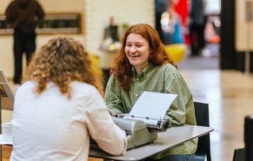 Back of the head of a man shot of a man and lady sitting at a desk with typewriter in front of him.