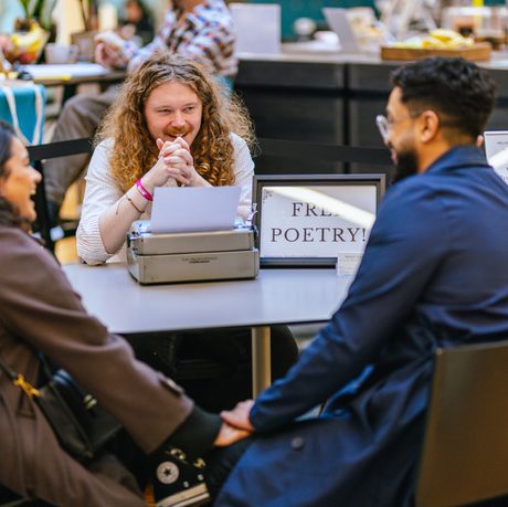 Two people sitting at a desk in front of a man with typewriter and sign that reads Free Poetry!