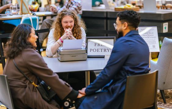 Two people sitting at a desk in front of a man with typewriter and sign that reads Free Poetry!