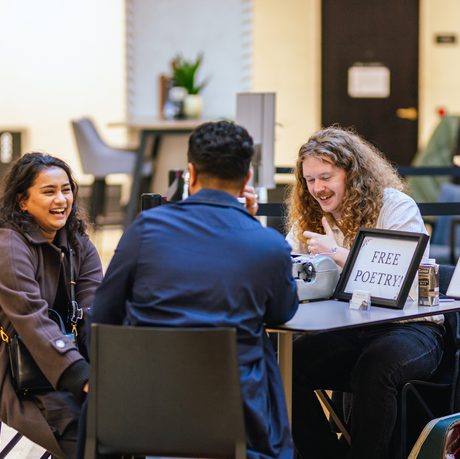 Two people sitting at a desk in front of a man with typewriter and sign that reads Free Poetry!