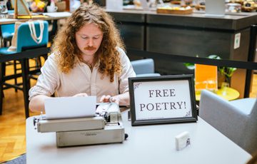 A man sitting at a desk with typewriter and sign that reads Free Poetry!