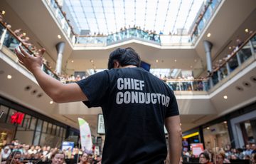 Back of a chief music conductor standing in front of a large crowd who are watching from three levels in a shopping centre.