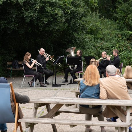 A group of musicians perform in front of a small crowd on benches in an outdoor setting.