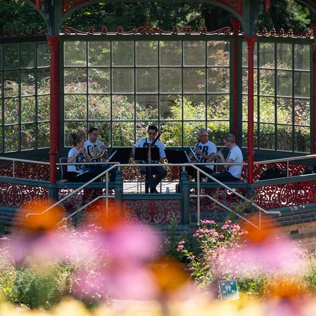 A group of musicians perform under an ornate bandstand in a garden.