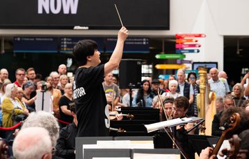 A music conductor in the centre of musicians, a crowd is watching in the background.