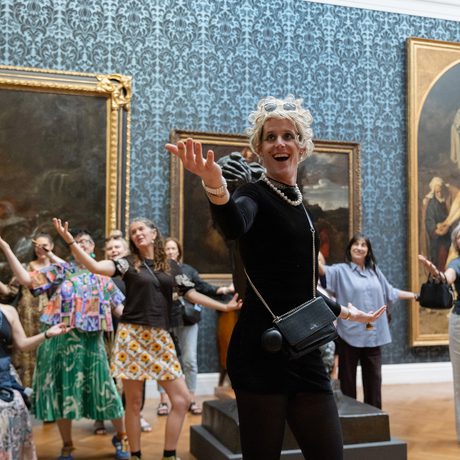 A person performing and holding their arm out, in a gallery setting with people behind copying the movement.