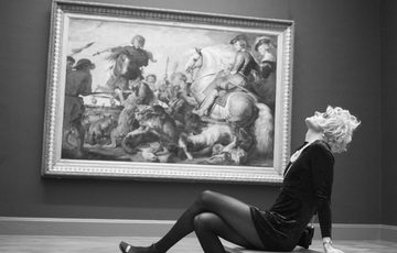 Black and white photo of a person leaning back on a bench in an art gallery with painting in the background.