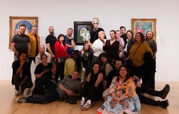 A group of people standing and sitting on the floor posing in an art gallery.
