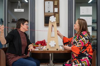 Two people sitting in front of a piece of equipment on a table in front and threading a red piece of wool.