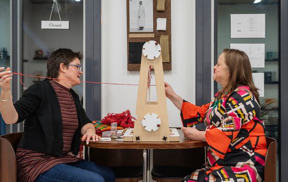 Two people sitting in front of a piece of equipment on a table in front and threading a red piece of wool.