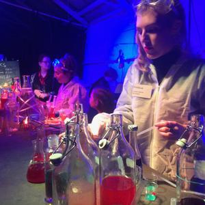 Two women are wearing white coats and have googles on their heads, above their eyes. There are bottles with different coloured liquid on the table in front of them.