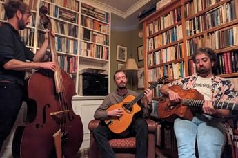 Three men playing music instruments including cello and guitar, in a room surrounded by books on shelves.