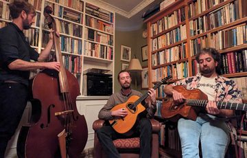 Three men playing music instruments including cello and guitar, in a room surrounded by books on shelves.