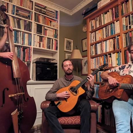 Three men playing music instruments including cello and guitar, in a room surrounded by books on shelves.