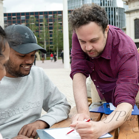 A man in purple shirt is sketching a picture on paper looked on by two other people