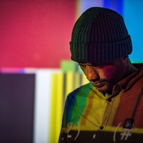 Antonio Roberts wearing a hat looking down, a colourful screen projected over the top.