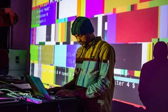 Antonio Roberts wearing a hat looking down at his computer, a colourful screen projected over the top.