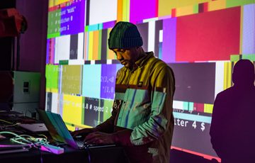 Antonio Roberts wearing a hat looking down at his computer, a colourful screen projected over the top.