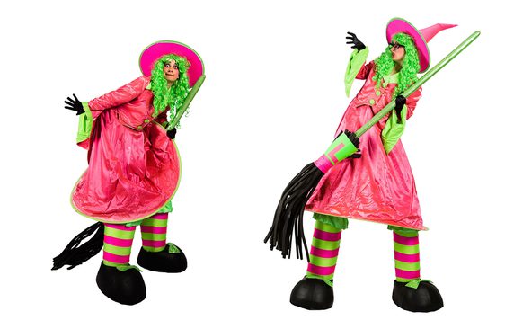 Two people on stilts, dressed in bright pink and green witch costumes with brooms.