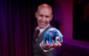 A smiling person holding a small toy elephant to the camera.