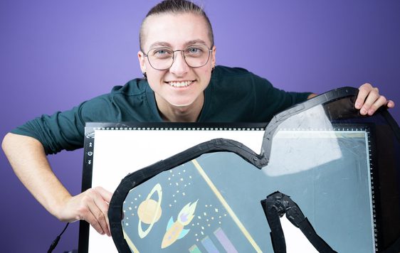 A smiling person holding a large pair of glasses in front of some paper.