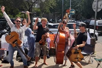 A group of musicians outside on a street, smiling with their hands in the air and holding a variety of string instruments.