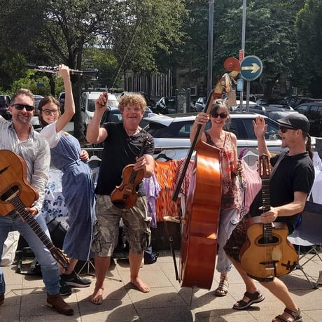 A group of musicians outside on a street, smiling with their hands in the air and holding a variety of string instruments.