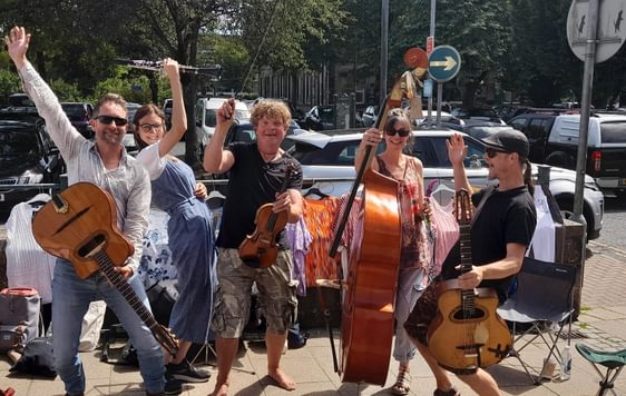 A group of musicians outside on a street, smiling with their hands in the air and holding a variety of string instruments.