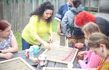 A group of people who are seated at a table are being helped by an artist with wool and weaving craft activity.