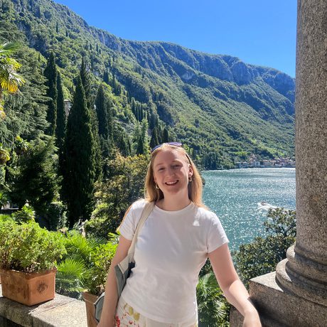 Photo of a lady wearing white and flowers, with a mountain landscape and lake in the background.