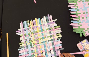 Arial shot of a table with hands weaving pastel coloured paper into a square.