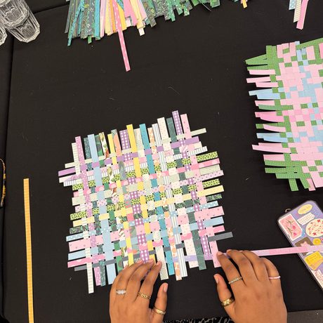 Arial shot of a table with hands weaving pastel coloured paper into a square.
