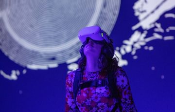 A person standing wearing VR headset and holding controllers, behind on the wall is a large graphic of an eye with a smiley face in the middle.