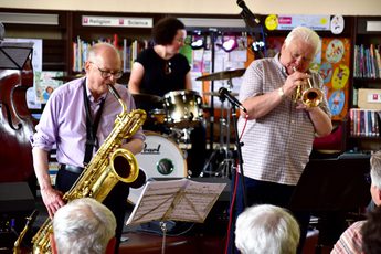 A group of people playing brass instruments in front of crowd in a library setting.