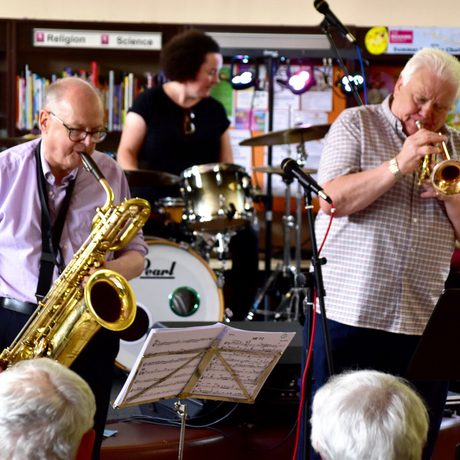 A group of people playing brass instruments in front of crowd in a library setting.