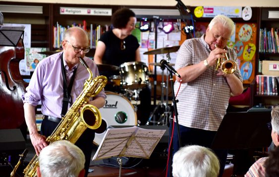 A group of people playing brass instruments in front of crowd in a library setting.