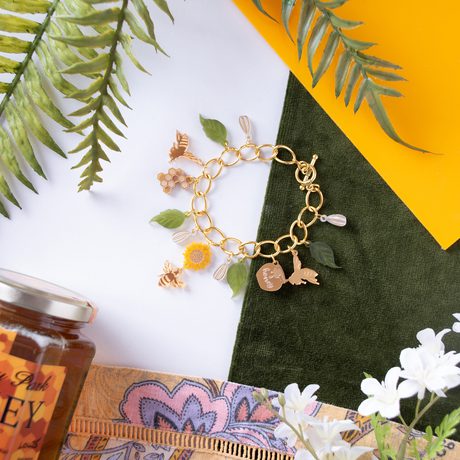 A gold bracelet with bee, leaf and sunflower design, laid out surrounded by leaves, fabric and honey pot.