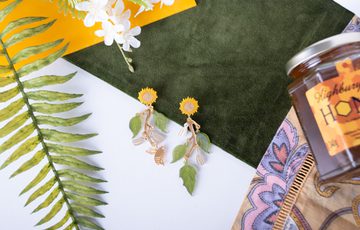 A set of earrings with sunflowers, leaf and bee design, laid out surrounded by leaves, fabric and honey pot.
