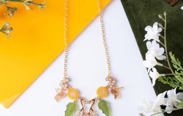 A necklace designed with leaves, sunflowers and bees, laid on a table surrounded by fabric, leaves and a honey pot.
