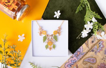 A necklace designed with leaves, sunflowers and bees in a box, surrounded by fabric, leaves and a honey pot.