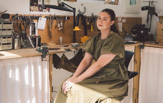 A person smiling sitting in a jewellery making workshop.