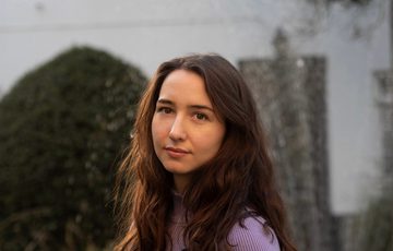 Head shot of a person with long brown hair wearing a lavender top outside of a white building and plants.