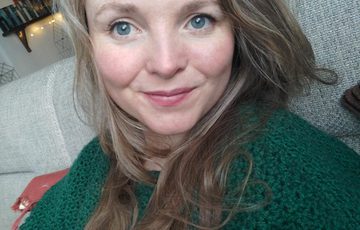 Image of a lady with blue eyes and long fair hair wearing a green top, sat on a grey sofa