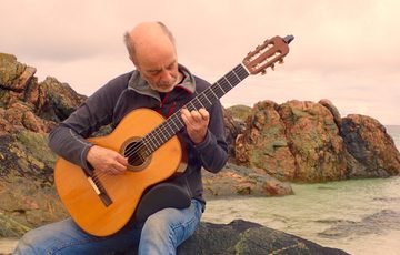 A man playing an acoustic guitar outside, a rocky landscape is in the background.